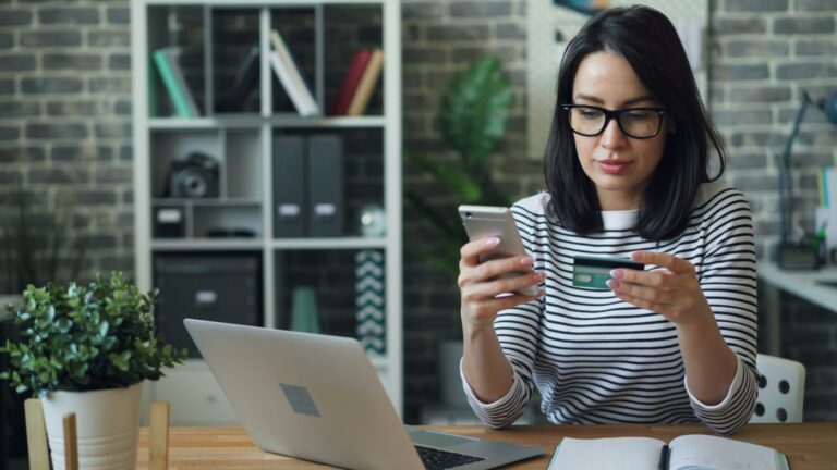 woman holding a bank card and typing into her phone with laptop on the desk
