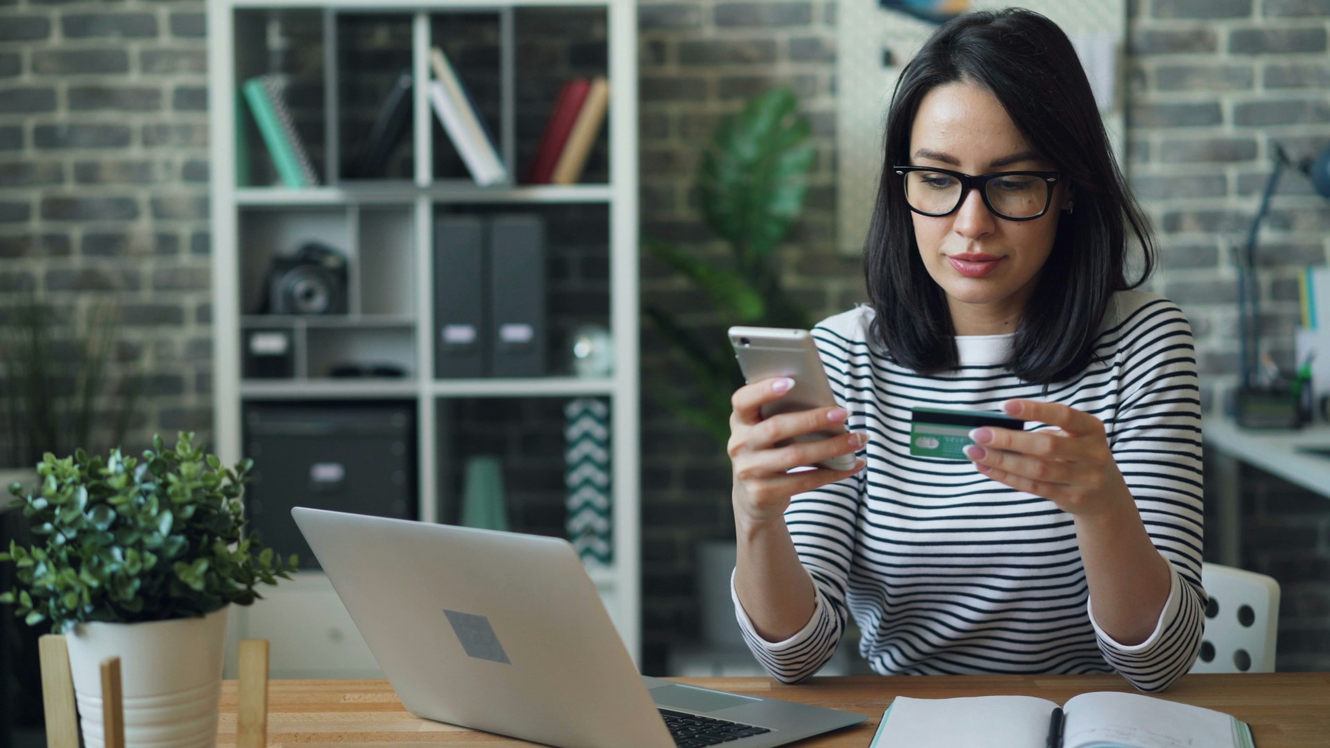 woman holding a bank card and typing into her phone with laptop on the desk