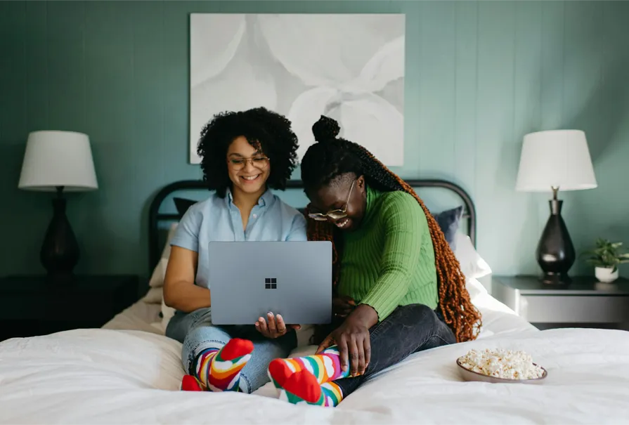 two people sitting on a bed looking at a laptop and laughing