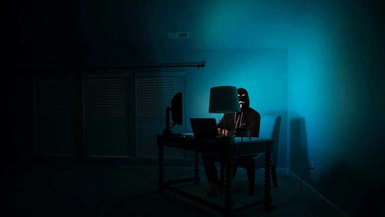 masked figure sits at a computer in the dark